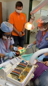 Dental team of three clinicians (two seated, one standing) performing procedure on patient; shade guide and instruments visible
